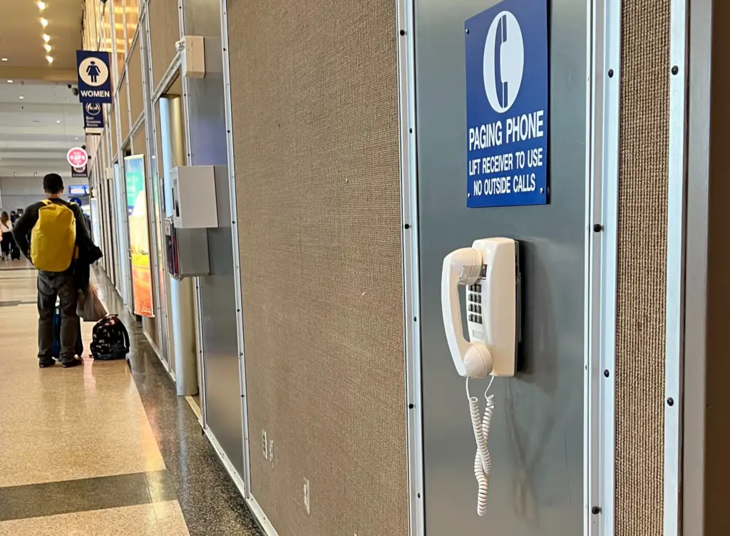 Wall-mounted paging phone in an airport hallway with a blue sign reading 'Paging Phone – Lift receiver to use, no outside calls,' and travelers walking in the background.